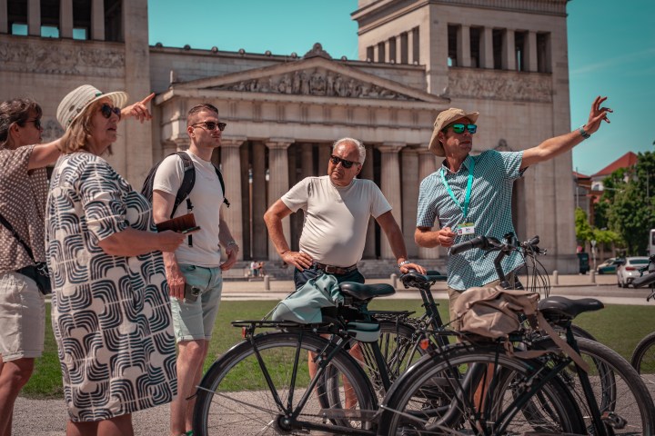 Group of people on a bicycle tour in front of a neoclassical building under a clear sky.