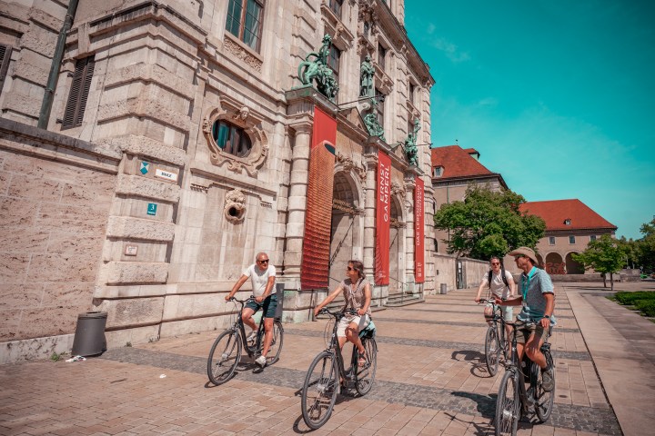 Four people on bicycles ride past a historic building under a clear blue sky.