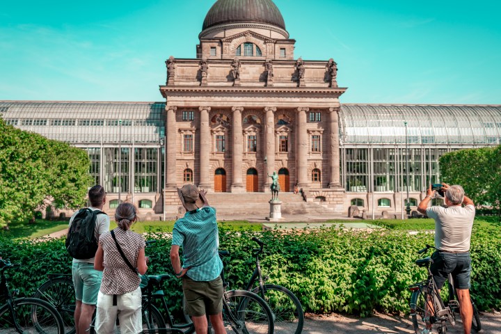 Tourists take photos of a historic building with bicycles in the foreground and trees on a sunny day.