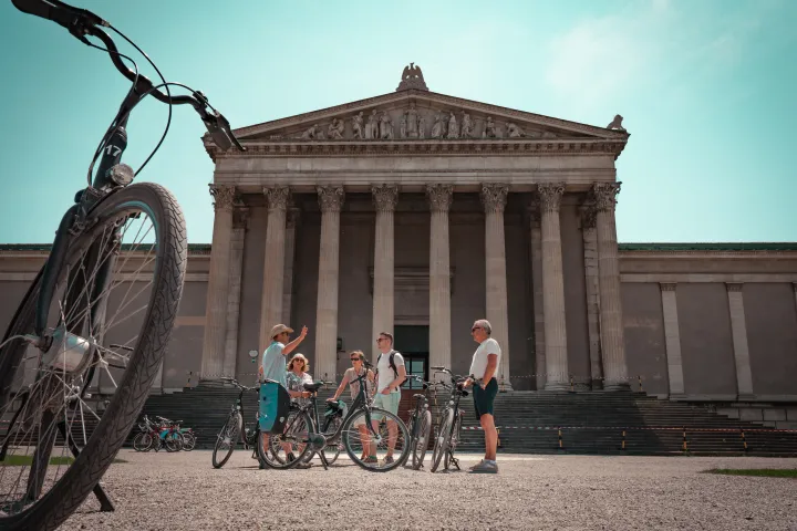 People with bicycles in front of a neoclassical building with columns.
