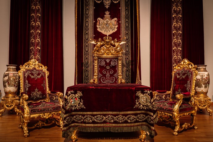 Ornate gold and red royal throne room with velvet drapes and tapestry.