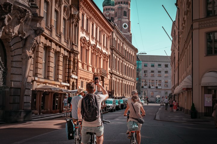 People cycling on a street between historic buildings with a cathedral in the background.
