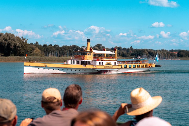 A vintage steamboat on a lake with people observing from the shore under a clear blue sky.