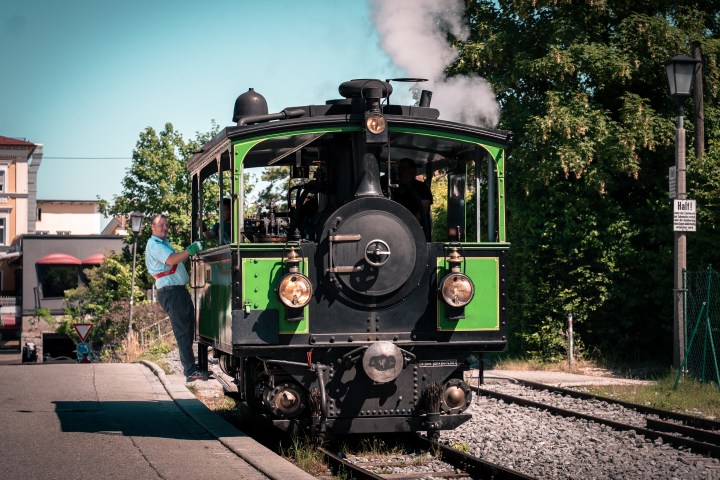 Person boarding a green steam locomotive on sunny day, surrounded by trees.
