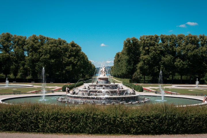 Ornate fountain with statues surrounded by circular gardens and trees under a clear blue sky.