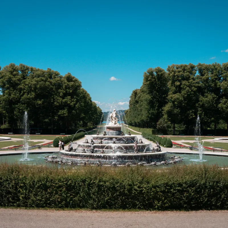 Ornate fountain with statues surrounded by circular gardens and trees under a clear blue sky.
