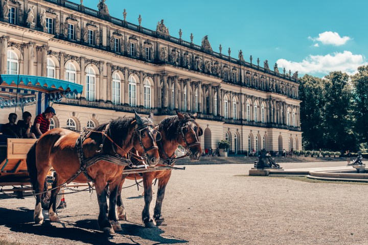 Two horses pull a carriage outside a grand historic building on a sunny day.