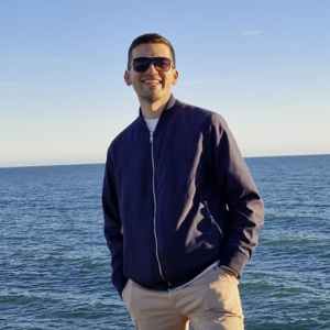 Person in sunglasses stands on rocky shore with ocean and clear sky in background.