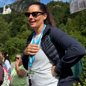 Person with sunglasses outdoors, wearing a lanyard, with a castle and mountains in the background.