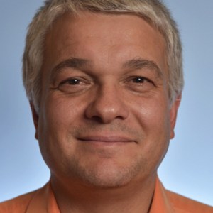 Smiling man with short gray hair in orange shirt on blue background.