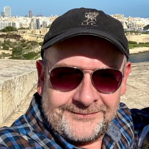 Man in sunglasses and cap taking selfie with cityscape in background.