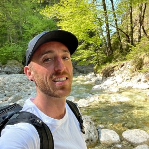 Person with backpack smiles beside a rocky creek in a wooded area.