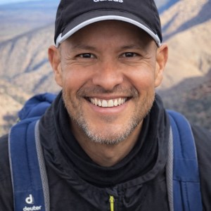 Smiling person wearing a cap and backpack, with mountains in the background.