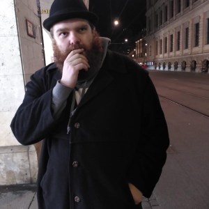 Bearded man in a black coat and hat standing on a dimly lit city street at night.