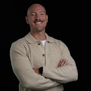 Smiling person with crossed arms wearing a beige sweater on a black background.