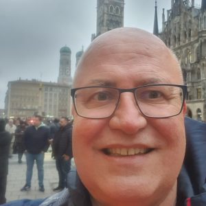 Smiling man with glasses in front of Gothic and domed tower buildings.