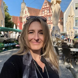 Woman smiling outdoors in a market square with European buildings in the background.