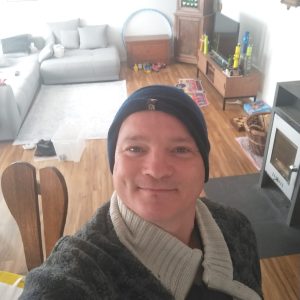 Person in beanie taking a selfie in a cozy living room with a gray couch and wooden floor.