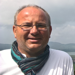 Smiling man with glasses and scarf outdoors with hilly background.
