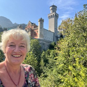 Smiling person in front of a large castle with trees and clear sky.