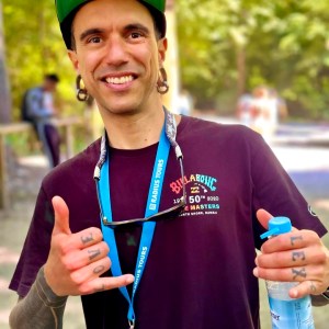 Smiling person with tattoos on fingers, wearing a cap, holding a water bottle and making a shaka sign.