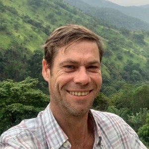 Smiling man outdoors with green hills and trees in the background.
