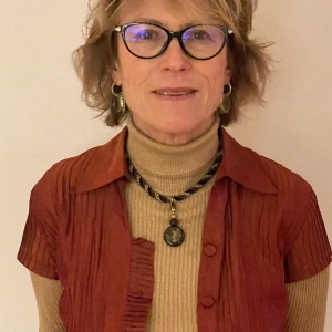 Woman with glasses, short hair, wearing an orange jacket and tan turtleneck, standing against a plain background.