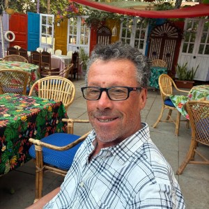 Man in glasses smiling, seated in a colorful outdoor cafe.