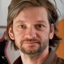 Man with medium-length hair and beard, wearing a brown shirt, looking into the camera.