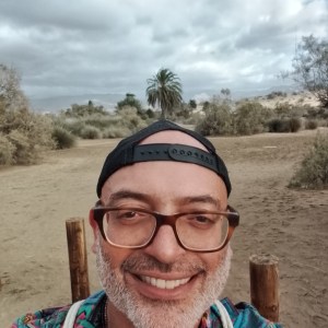 Man with glasses and cap smiling outdoors in desert landscape.