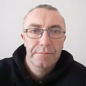 Person with glasses and short hair, wearing a black hoodie, looking directly at the camera against a plain background.