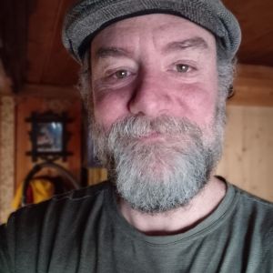 Man with a beard and flat cap smiling indoors with wooden background.