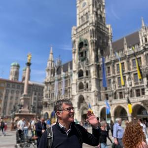 Smiling man waving in a busy square with a historic tower in the background.