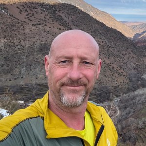 Bald man in yellow jacket smiling, with mountainous landscape in the background.