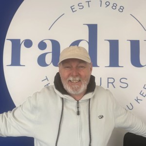 Smiling man in white hoodie and cap stands in front of a large sign with 'radius' partially visible.