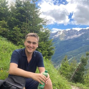 Smiling person holding a bottle, sitting on grassy hillside with trees and mountains in the background.