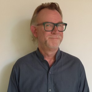 Man with glasses smiling, wearing a dark gray shirt against a plain background.