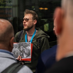 Man with glasses holding photo, speaking to a group of people outdoors.