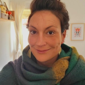 Person wearing a green and yellow scarf, smiling indoors with light background.