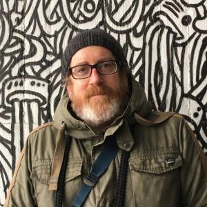 Person in beanie and glasses stands in front of black and white graffiti wall.