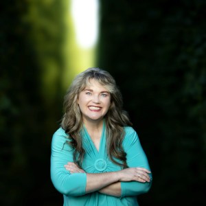 Smiling person with long hair in teal shirt, arms crossed, green blurred background.