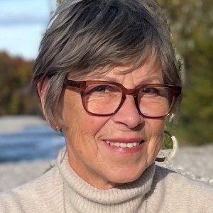 Smiling woman with short gray hair and glasses in a cream sweater outdoors.