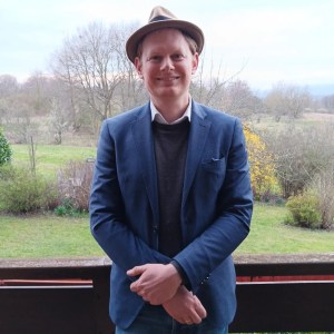 Man in hat and blue blazer standing in front of a scenic landscape.